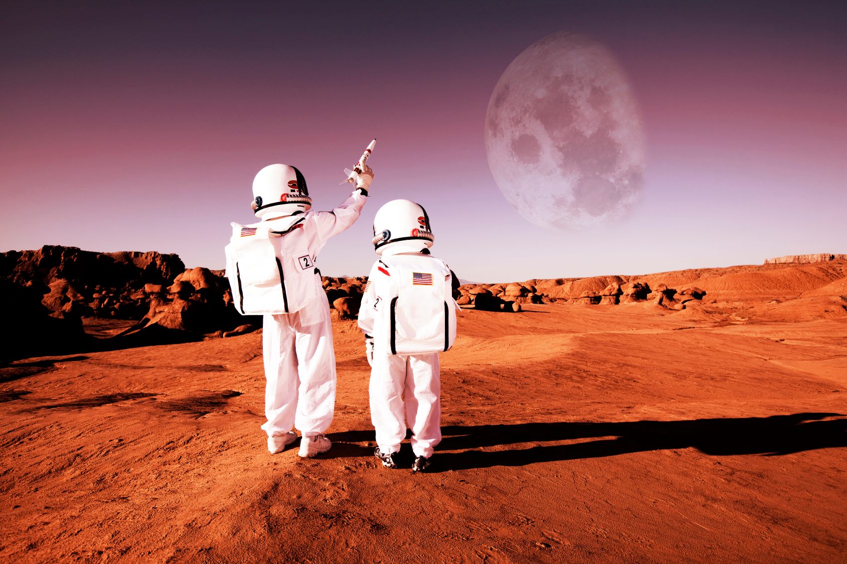 Two young astronauts on a red landscape, one pointing a toy rocket towards a large moon